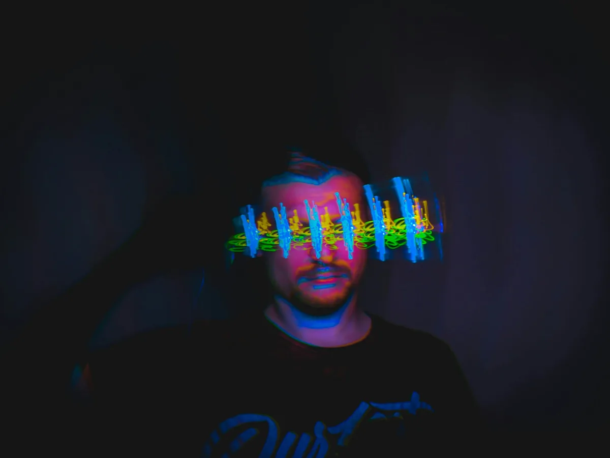 Man in a dark room with vibrant blue, green, and yellow light trails blurred across his eyes in a long-exposure effect