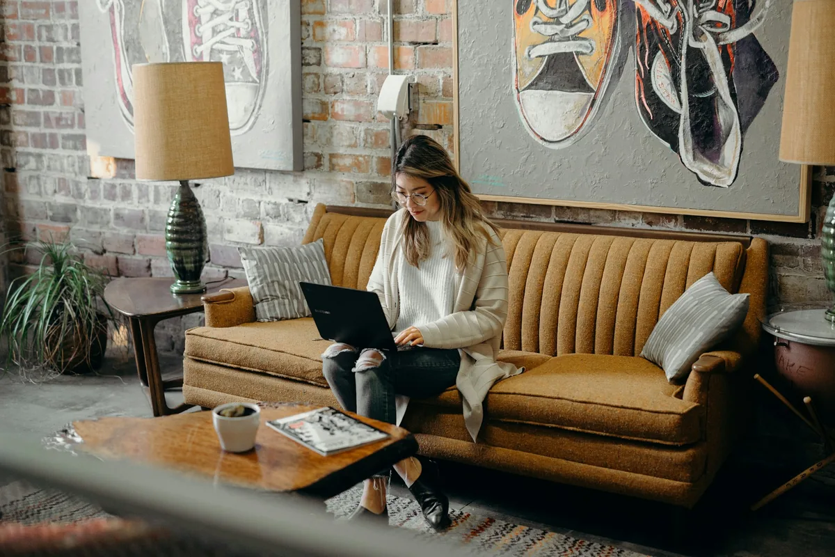 A woman working on her laptop while sitting on a mustard-yellow couch in a stylish, rustic room