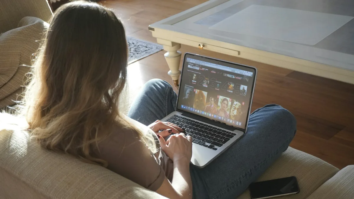 Over-the-shoulder view of a woman sitting on a couch, browsing Netflix on a laptop