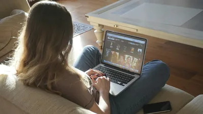 Over-the-shoulder view of a woman sitting on a couch, browsing Netflix on a laptop