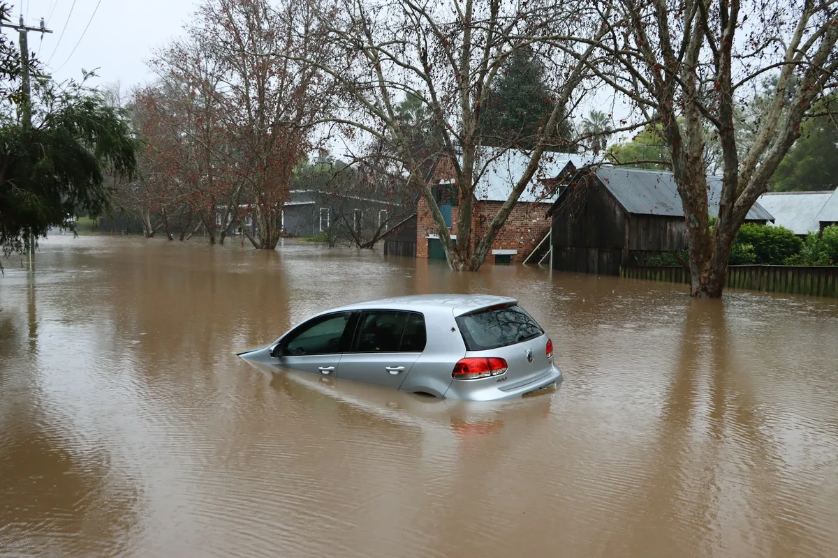 A silver hatchback partially submerged in deep, murky floodwaters on a residential street